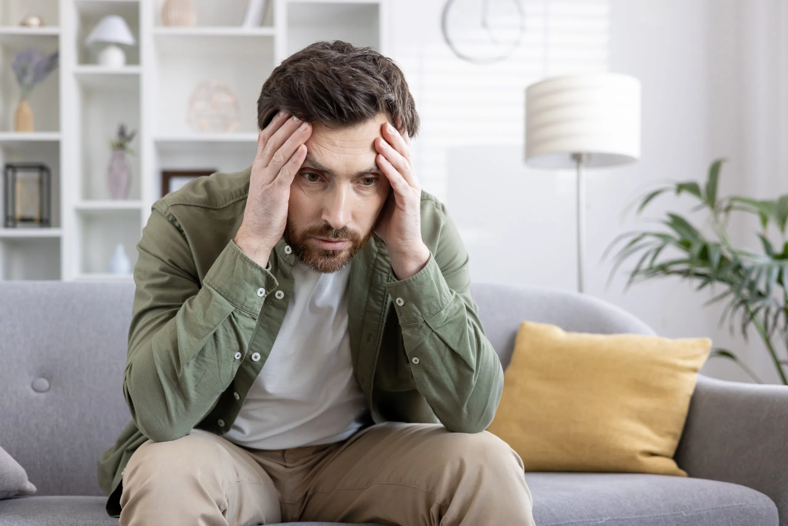 Man sitting on a sofa holding his head with both hands, looking stressed and overwhelmed