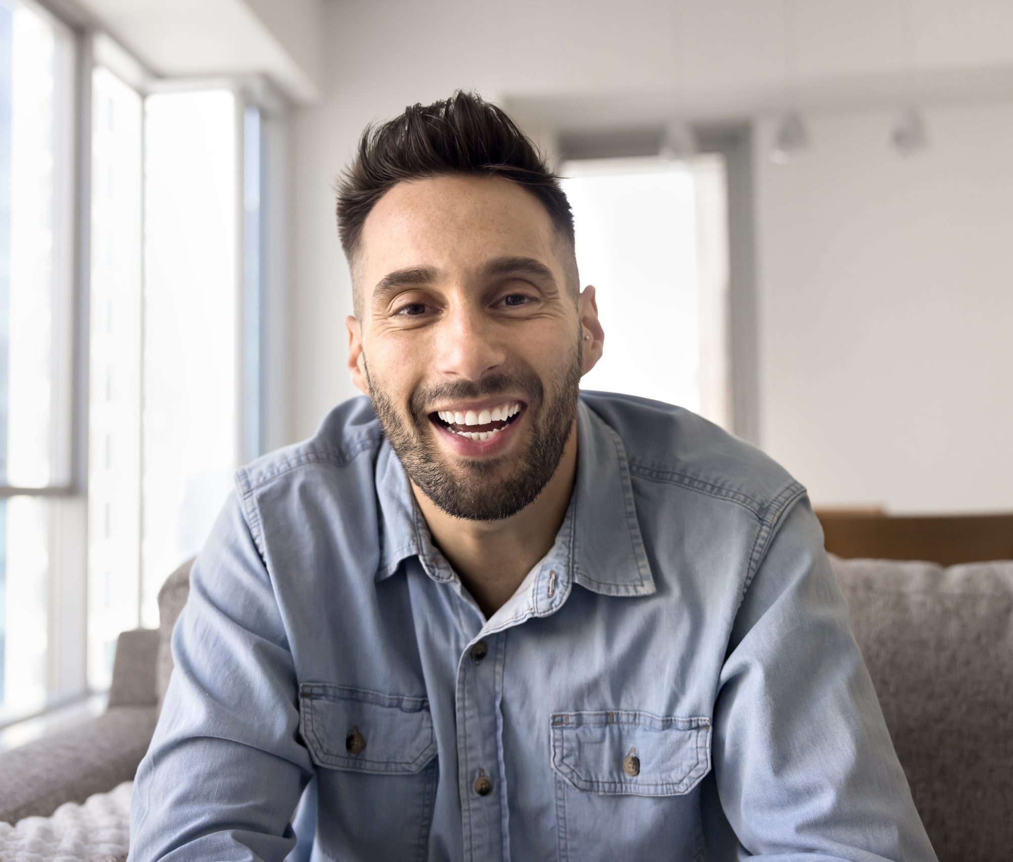 Smiling man in a light blue shirt sitting on a couch near bright windows
