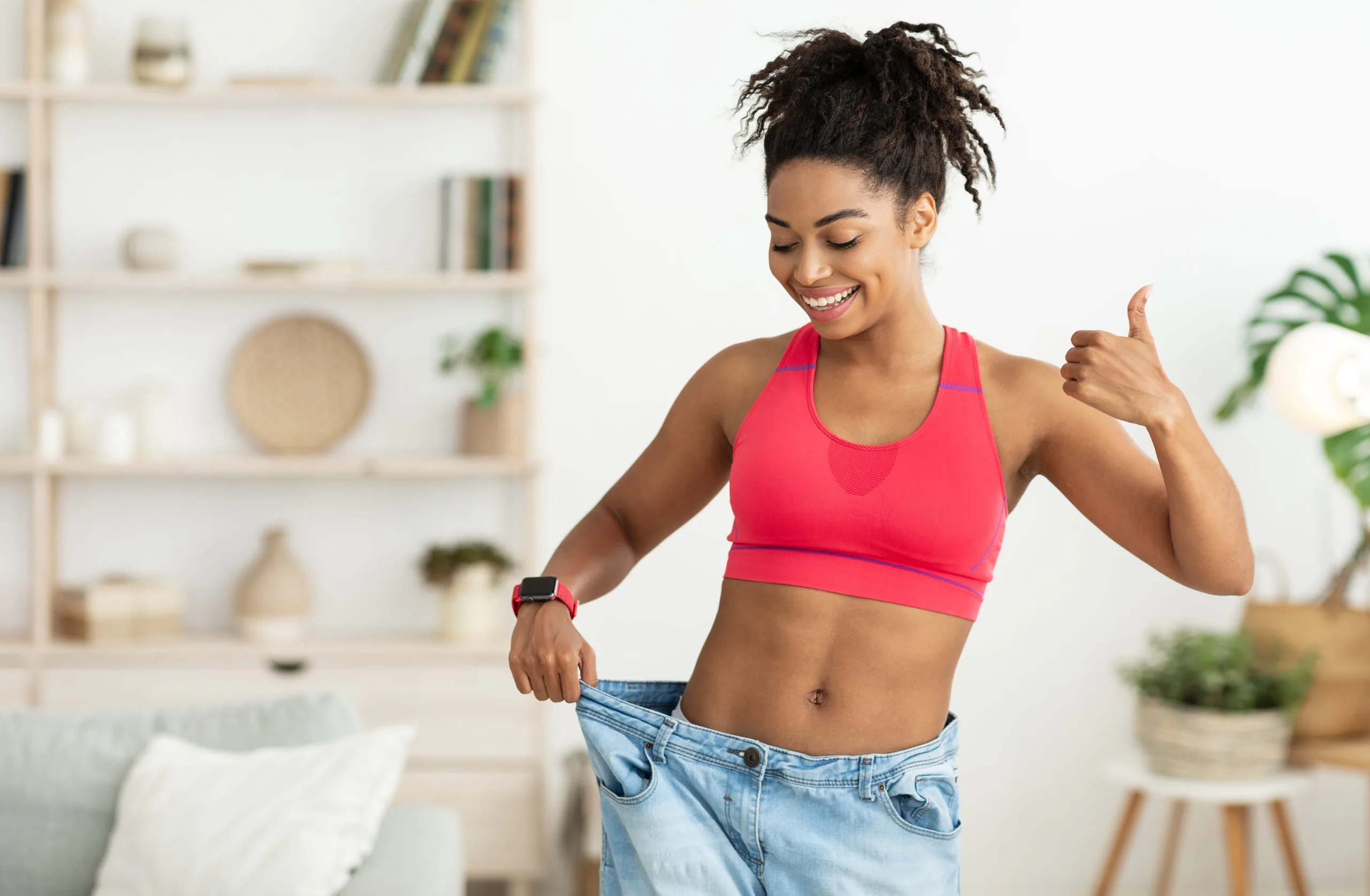 Woman in pink sports bra giving thumbs up while holding oversized jeans at waist