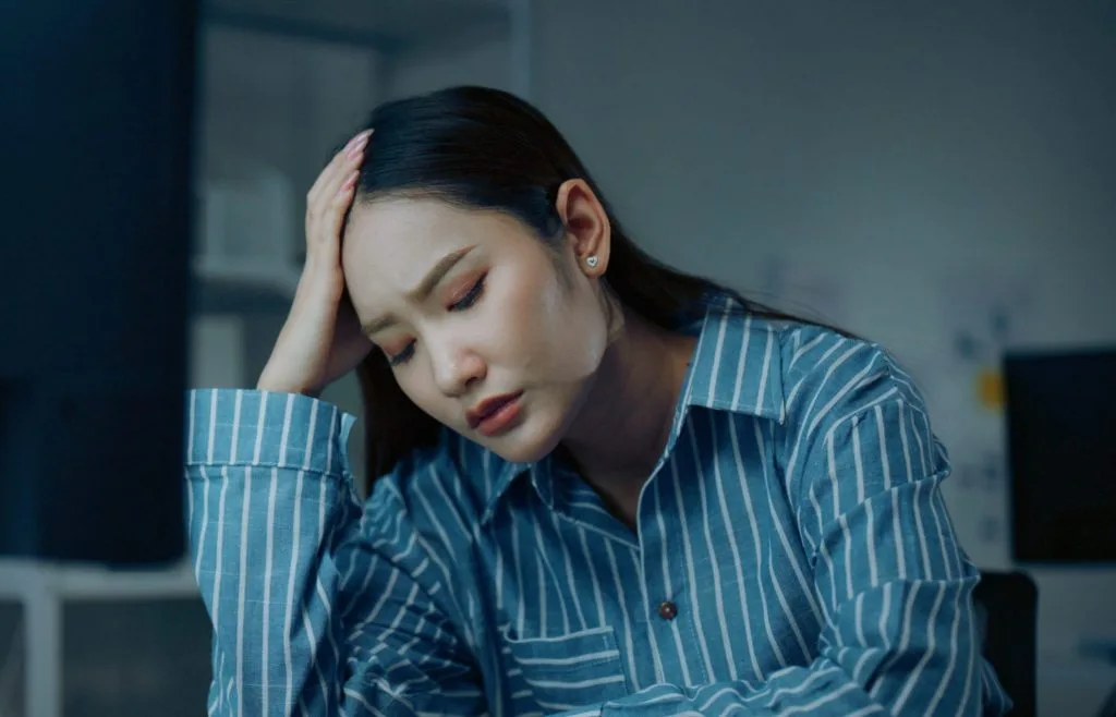 Woman in blue striped shirt holding her head while working at a desk, appearing stressed and fatigued