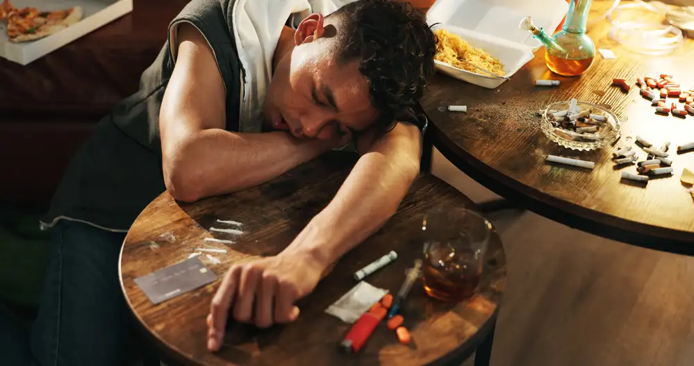 Person slumped over table surrounded by pills, cigarettes, and alcohol, depicting substance abuse