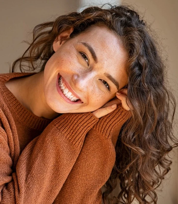 Smiling woman in her 30s in a warm rust sweater, resting her head on her arms