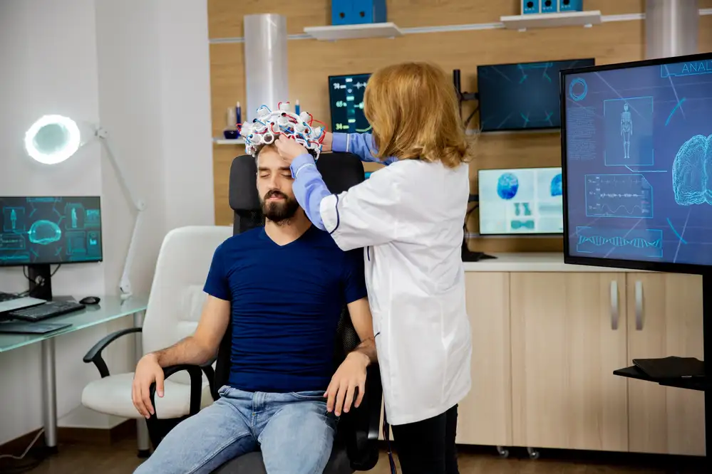 Healthcare provider placing a TMS coil headset on a patient's head in a modern clinical setting with brain imaging displays