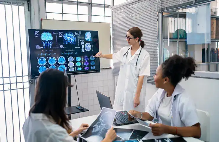 Clinician pointing to brain scan images on monitor while discussing results with two colleagues in a clinical setting