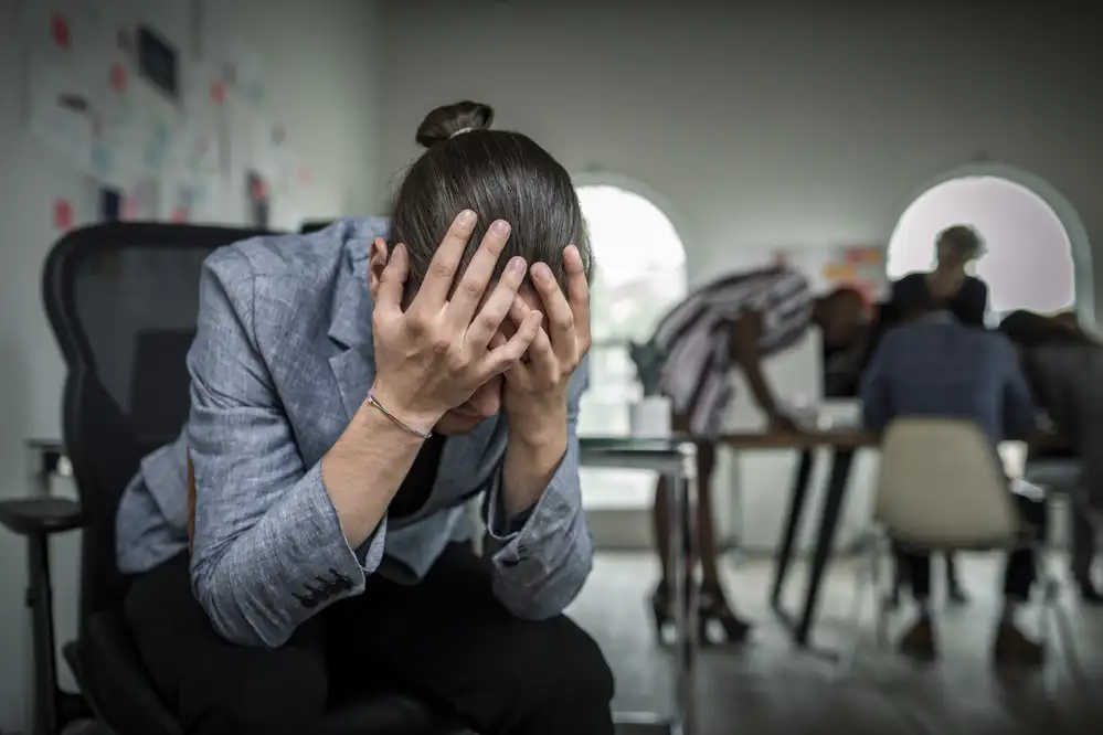 Person in business attire holding their head with both hands in a waiting room, showing signs of anxiety or stress