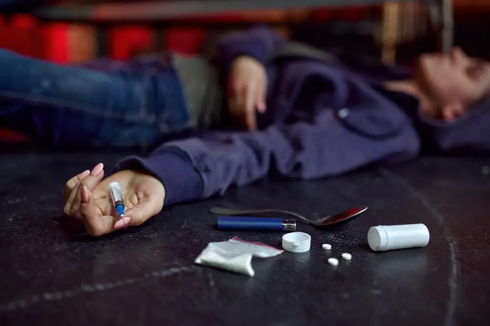 Person lying on floor holding syringe with drug paraphernalia and substances nearby