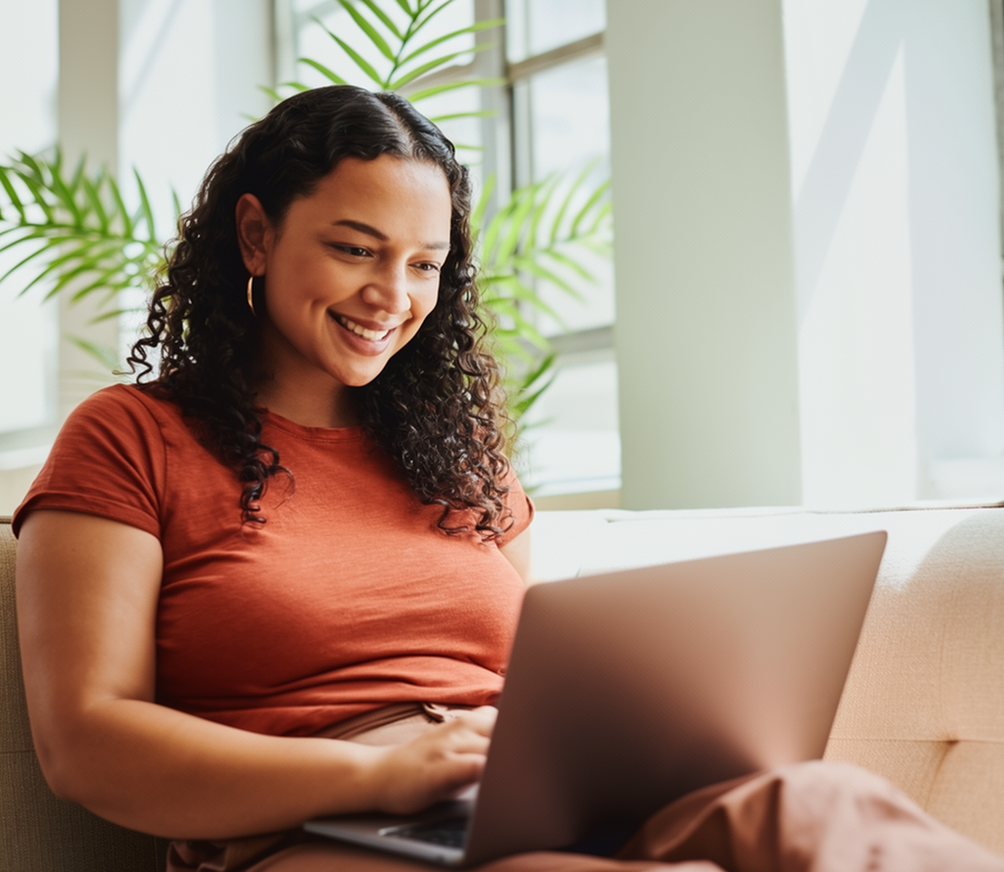 Woman in red shirt smiling while using laptop at home near window with plant