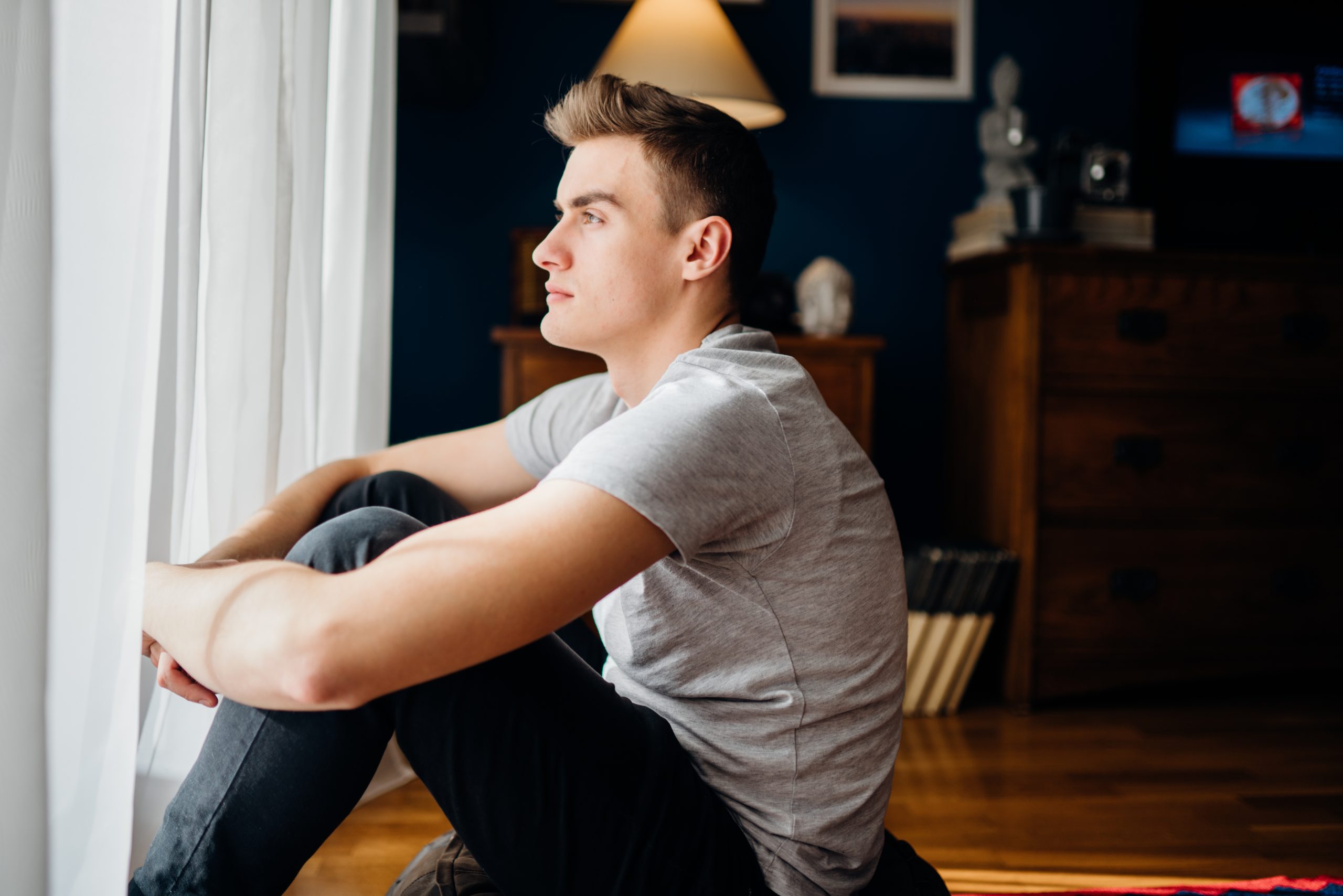 Young man sitting by window at night, looking out with contemplative expression
