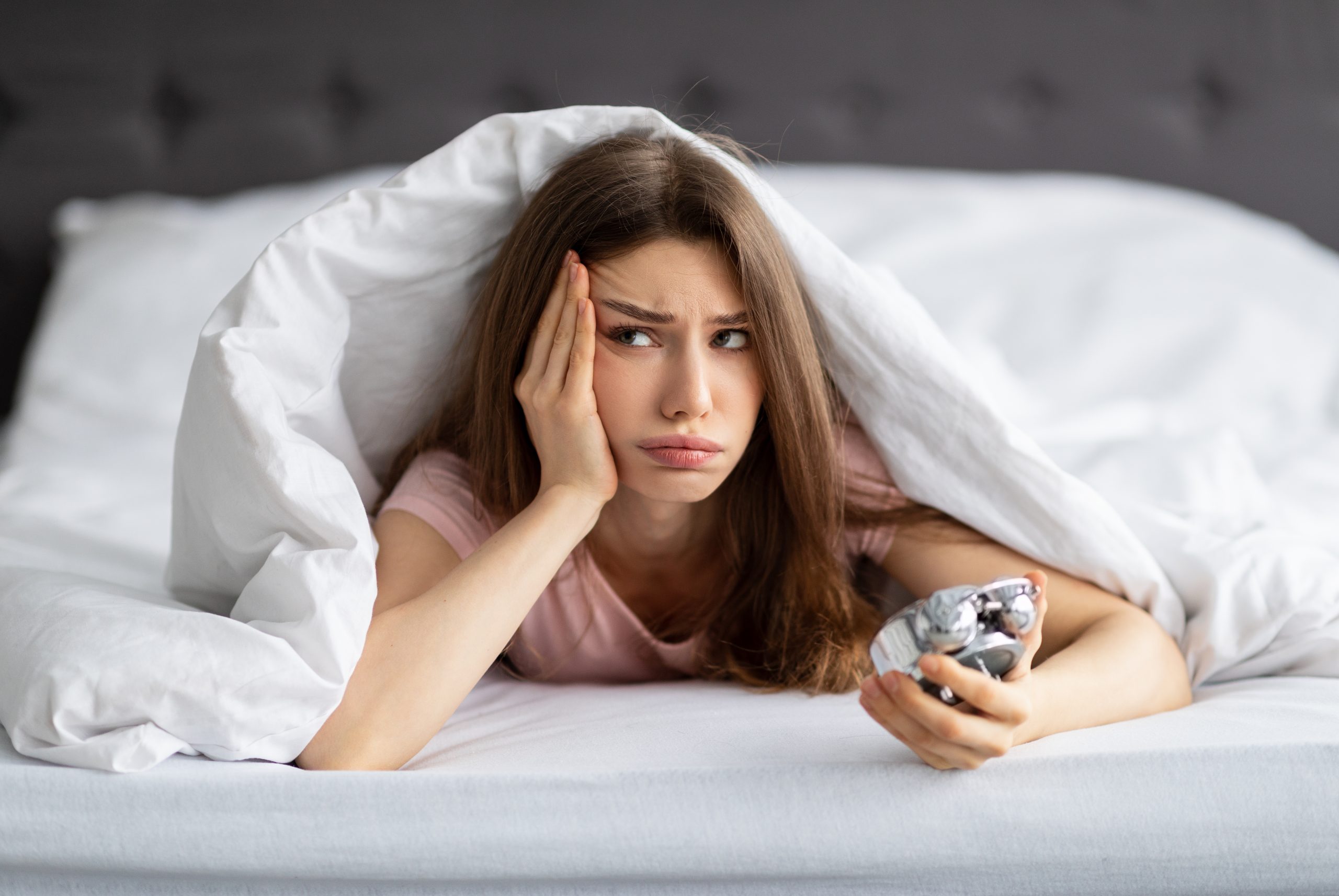 Woman under white blanket in bed holding a pill bottle, looking worried