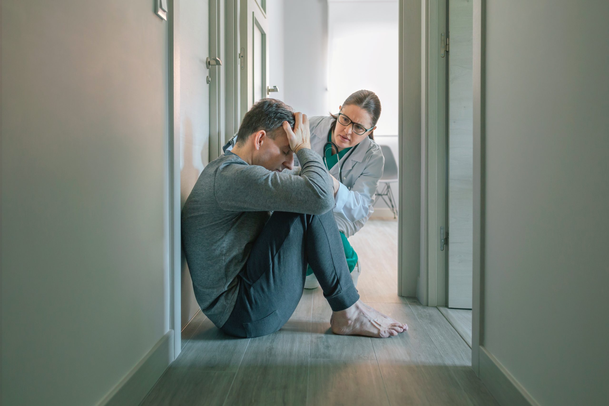 Healthcare provider in white coat and teal stethoscope speaking with distressed man sitting in hallway