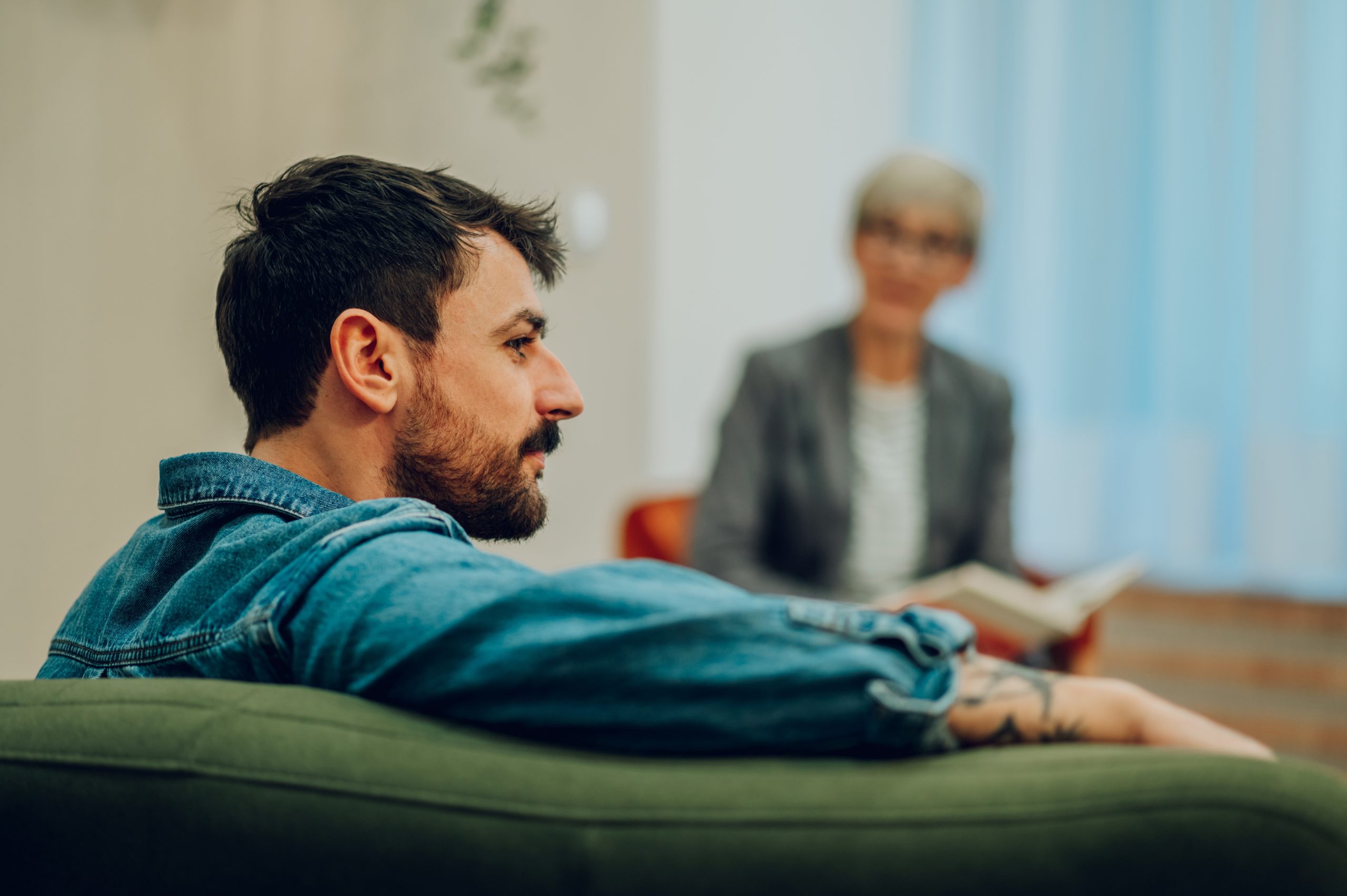 Man in denim shirt sitting on couch facing counselor in background