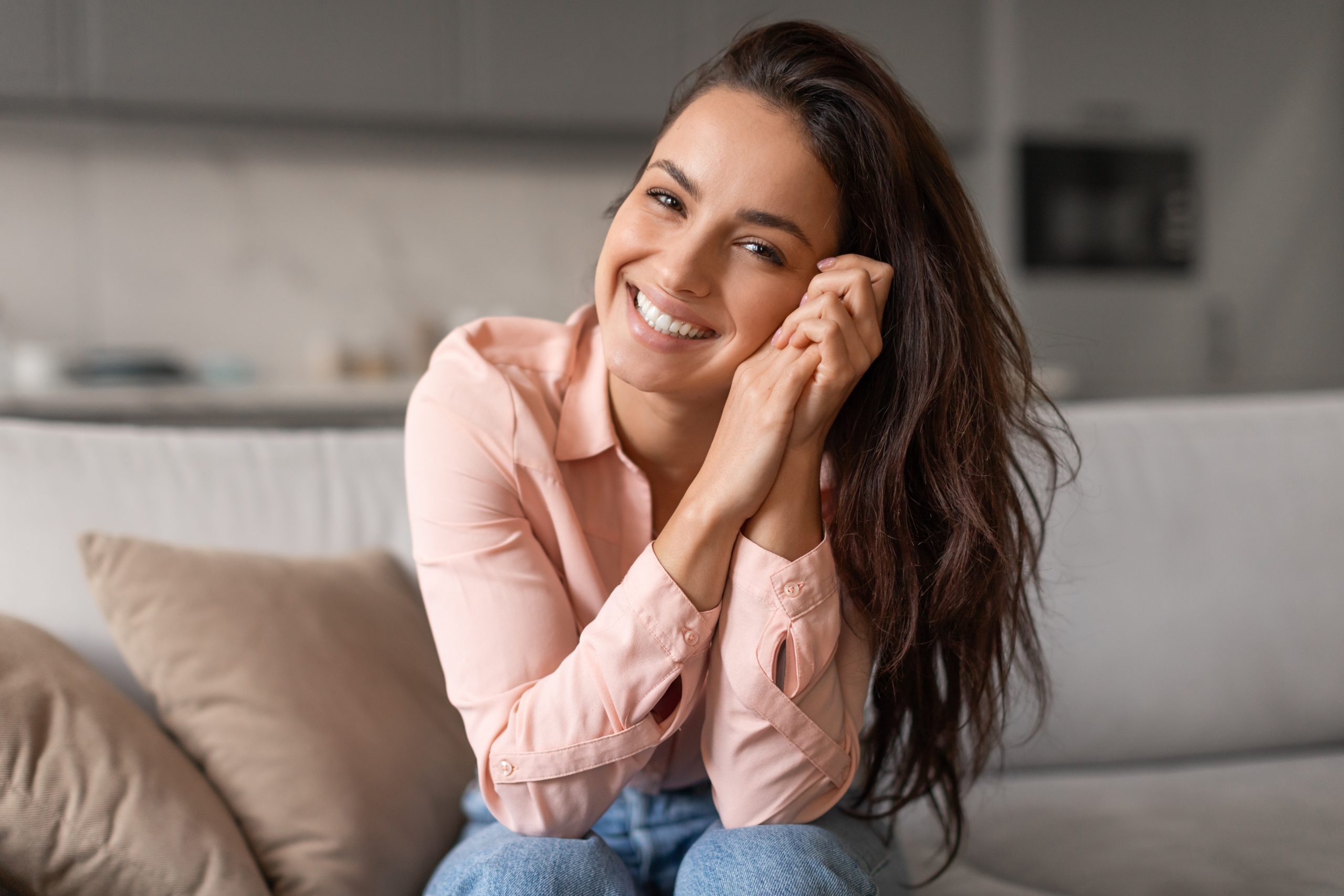 Woman in pink shirt smiling at camera while sitting on couch in modern home