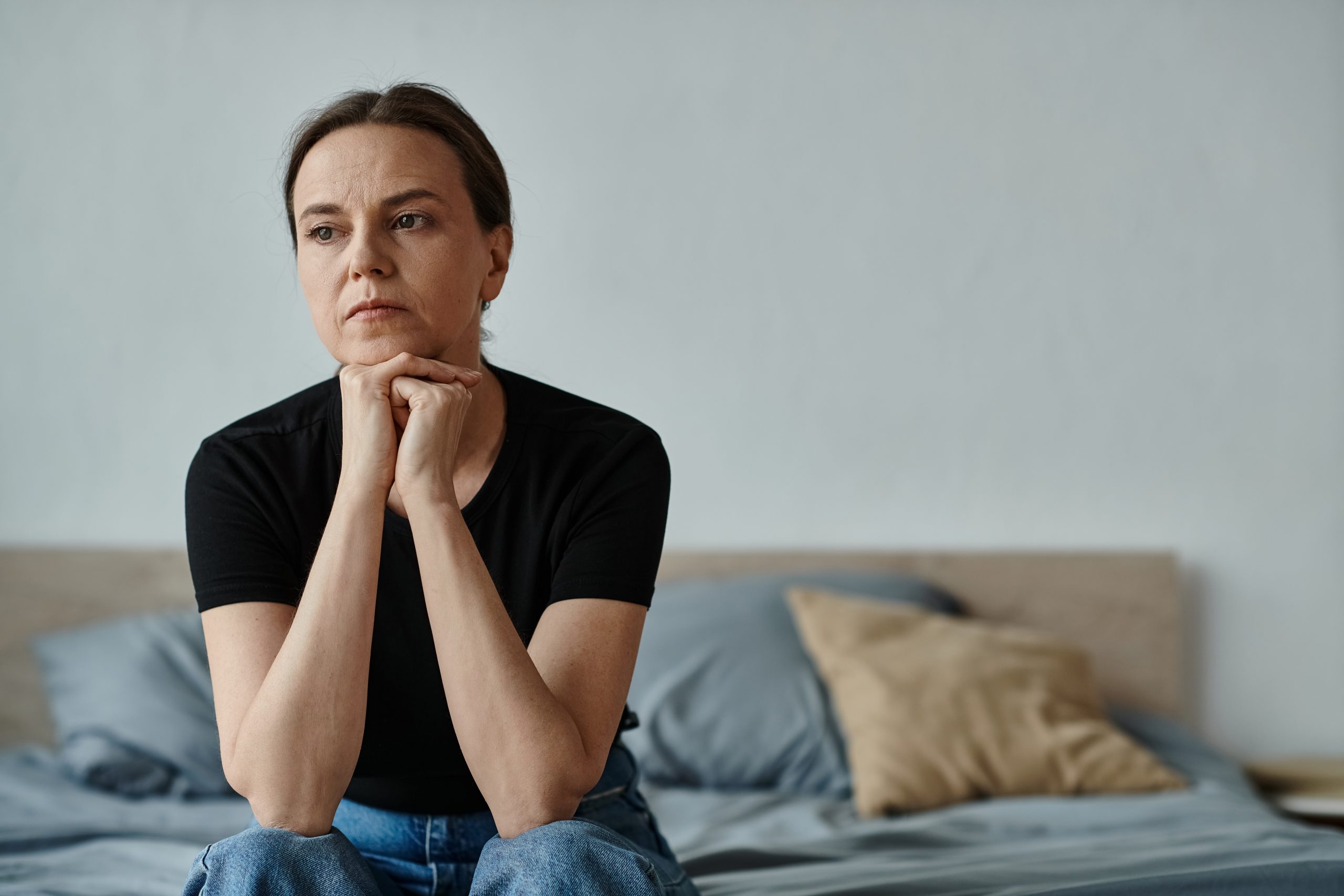 Woman in black shirt sitting on bed with hands clasped under chin, looking pensive