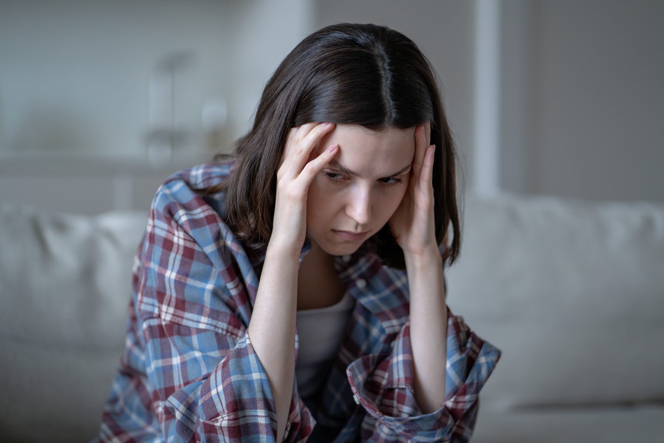 Woman in plaid shirt holding her head with both hands, looking distressed on a couch
