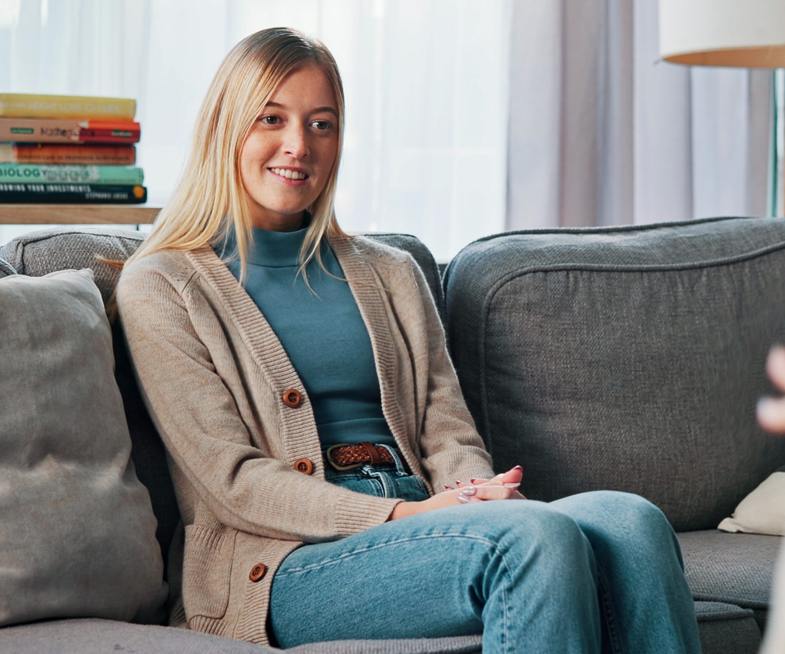 Woman sitting on couch smiling at camera in bright living room with books on shelf