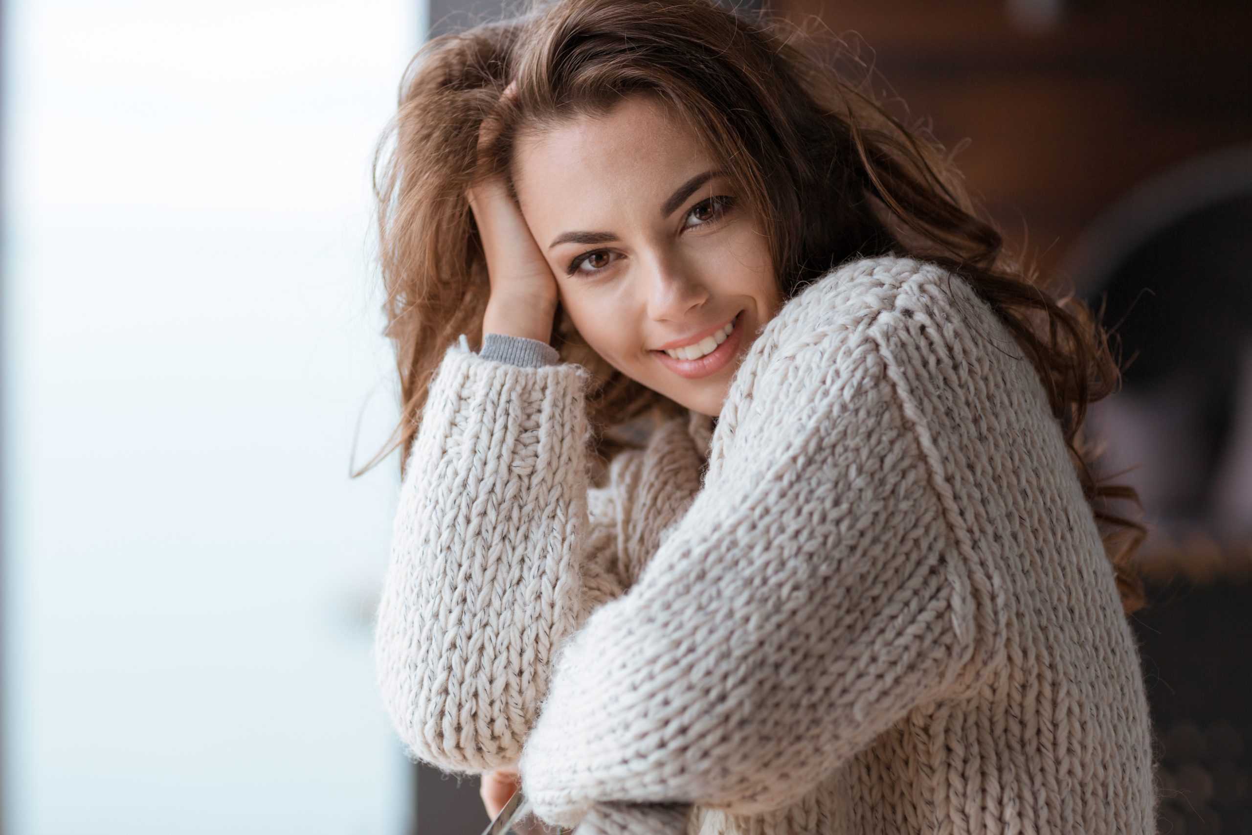 Woman in cream sweater smiling at camera with hand near face