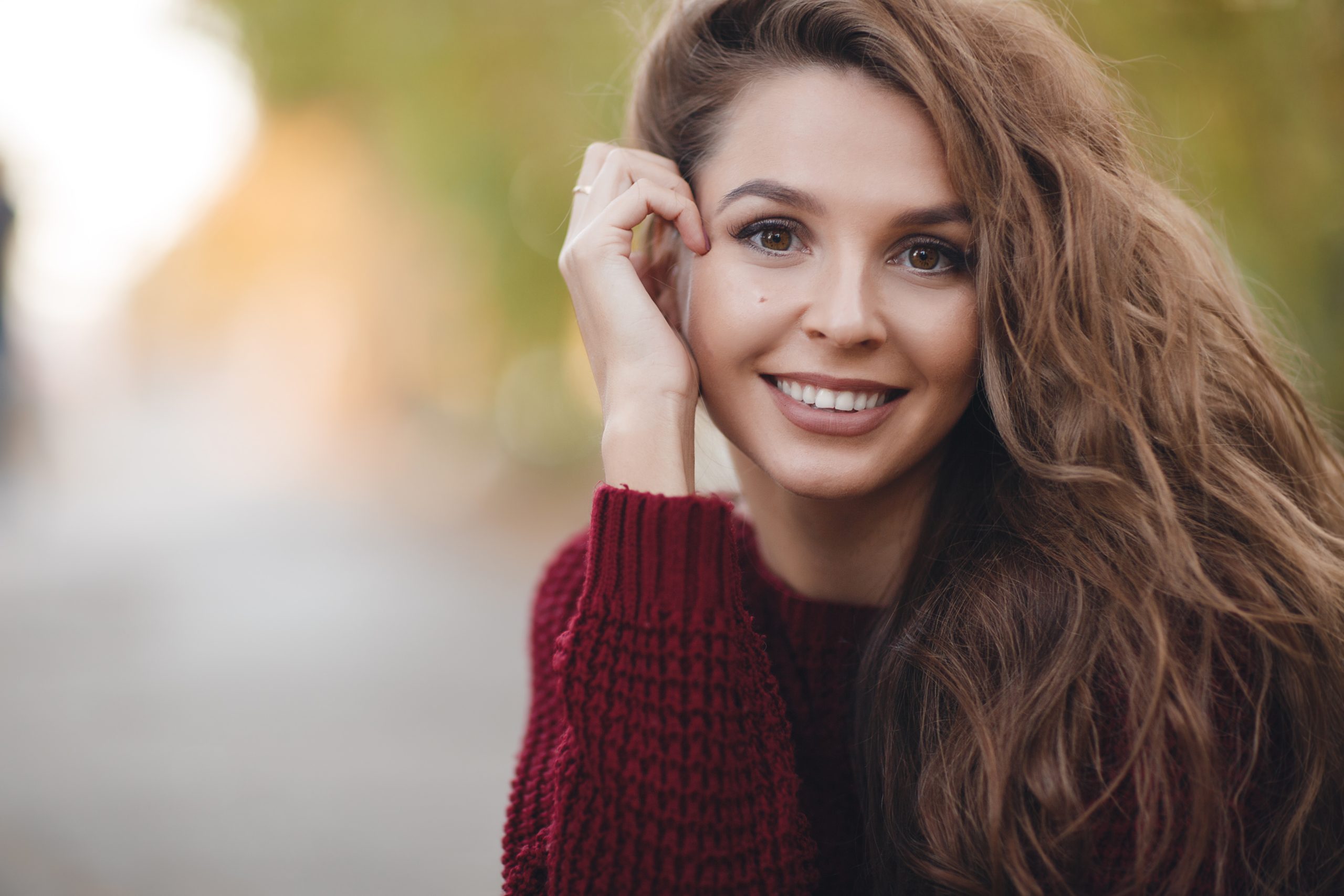 Woman in burgundy sweater smiling outdoors with hand near face