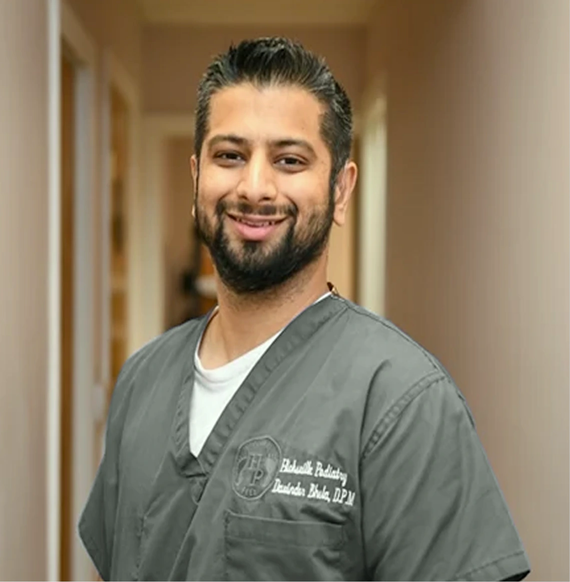 Dr. Bhela wearing gray medical scrubs with clinic logo, smiling in hallway