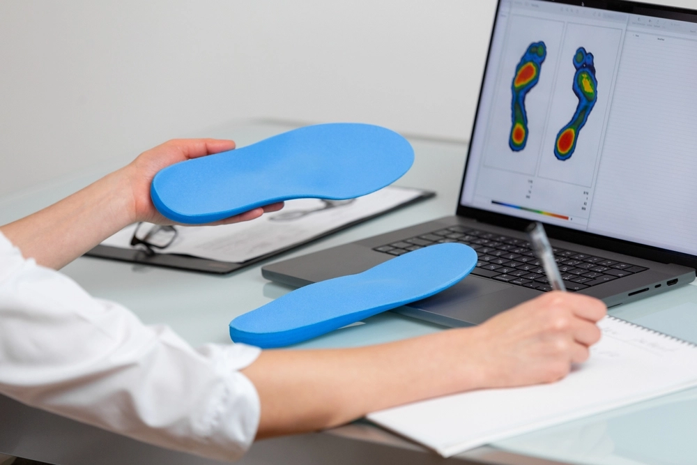 Podiatrist holding blue orthotic insoles beside laptop displaying thermal foot scan