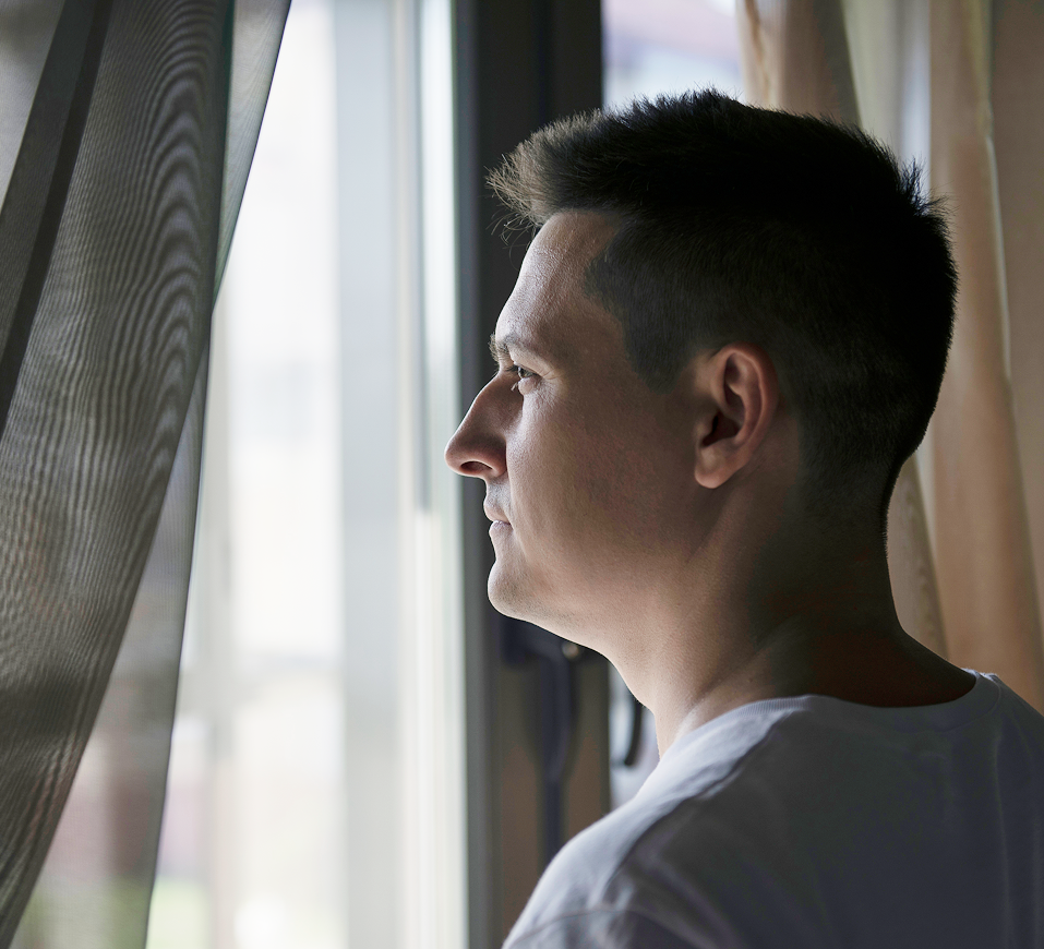 Man in profile gazing out a bright window with curtains