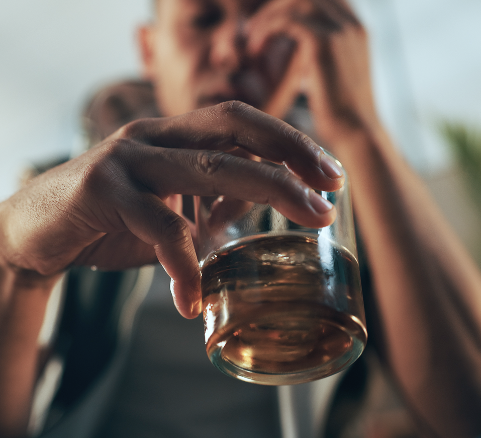 Person holding a glass of amber liquid with blurred figure in background
