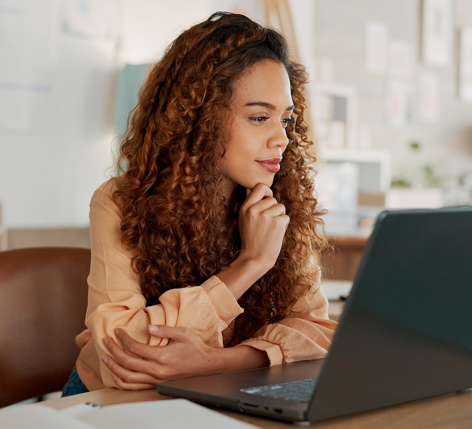 Woman with curly reddish-brown hair thinking while looking at a laptop in a bright office setting