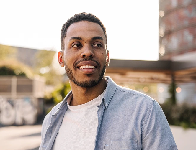 Man smiling outdoors wearing light blue shirt, seated in urban setting with brick building behind