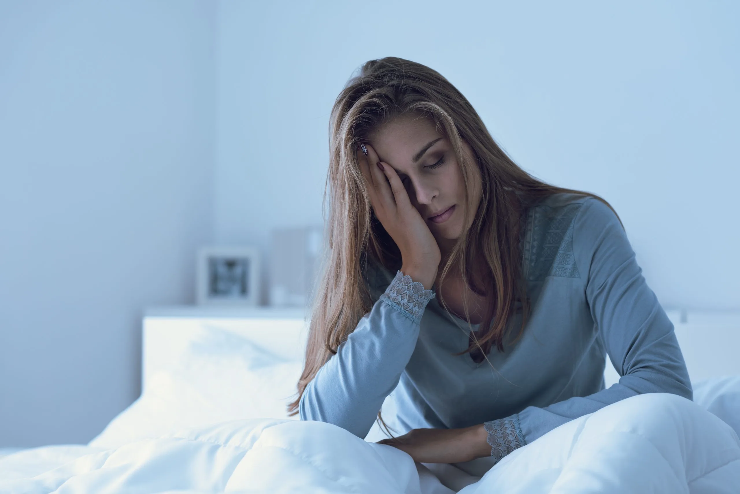 Woman in blue shirt sitting on bed with hand to face, appearing distressed