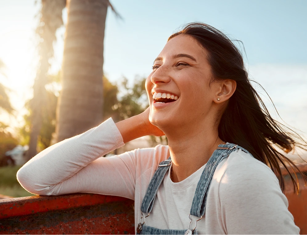 Woman smiling outdoors at sunset near a palm tree, wearing casual white and denim clothing
