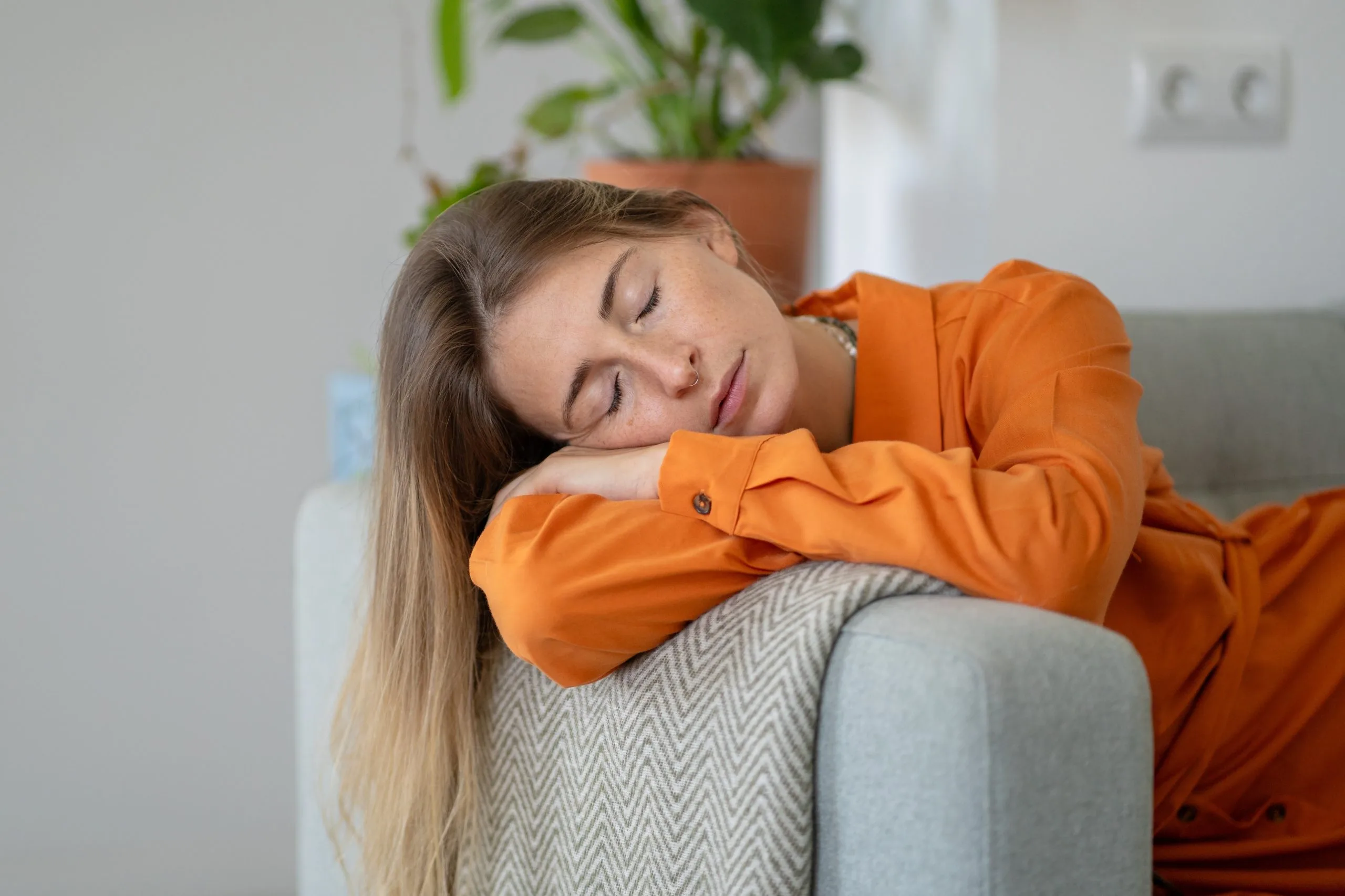 Woman in orange shirt resting head on couch pillow with eyes closed at home