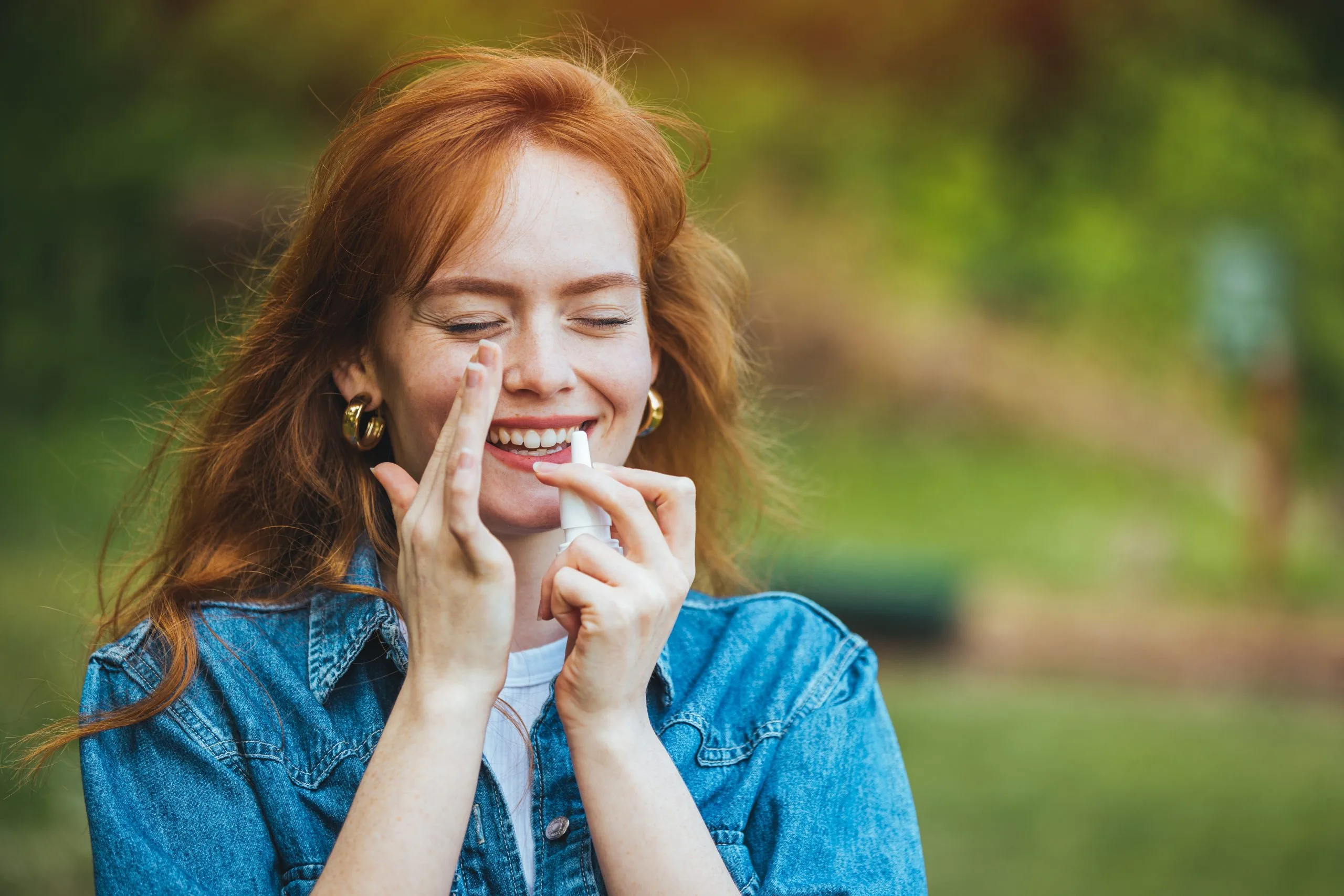 Smiling woman with red hair wearing denim shirt, hands near her face outdoors