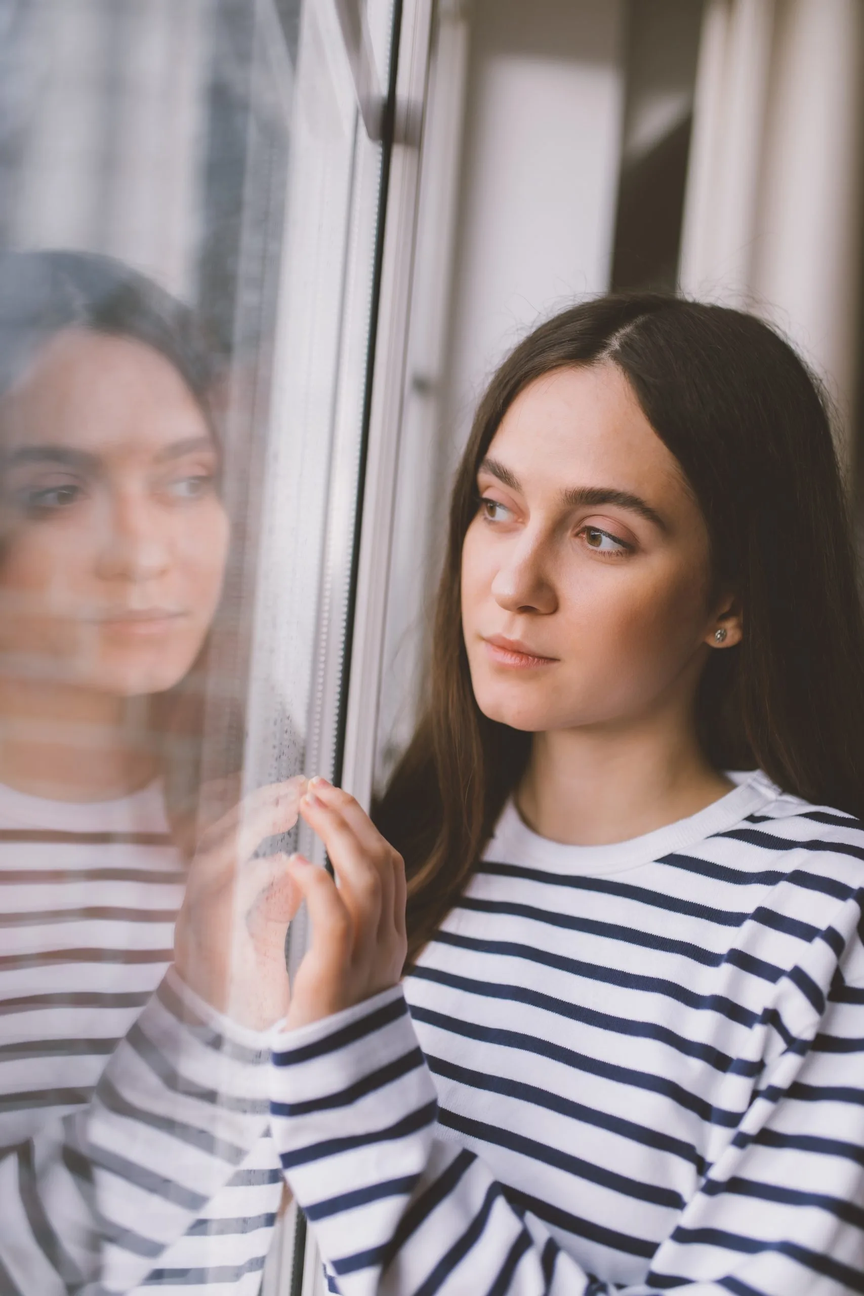 Woman in striped shirt gazing out window with reflection visible in glass