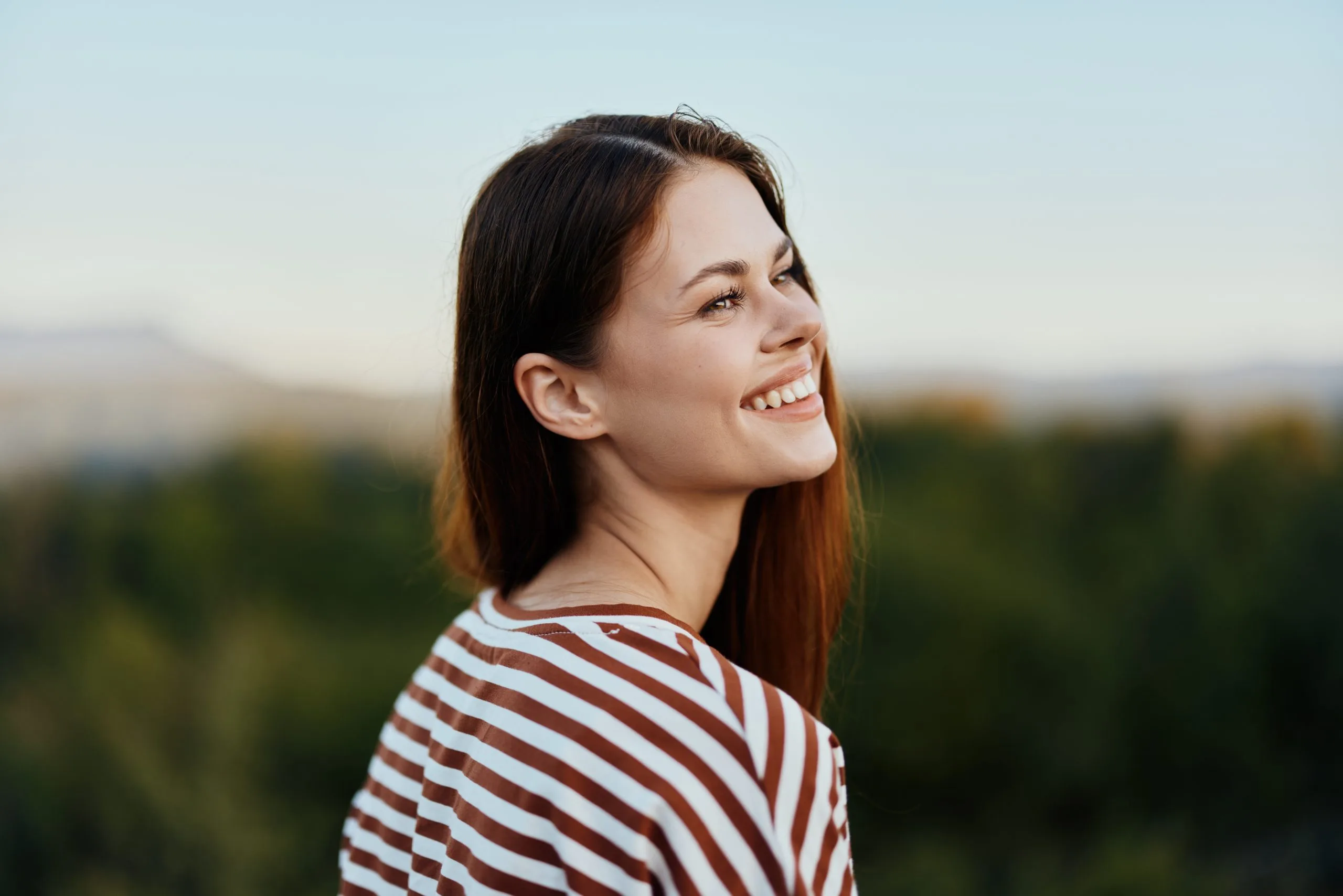 Woman smiling outdoors wearing a striped shirt with blurred trees in background