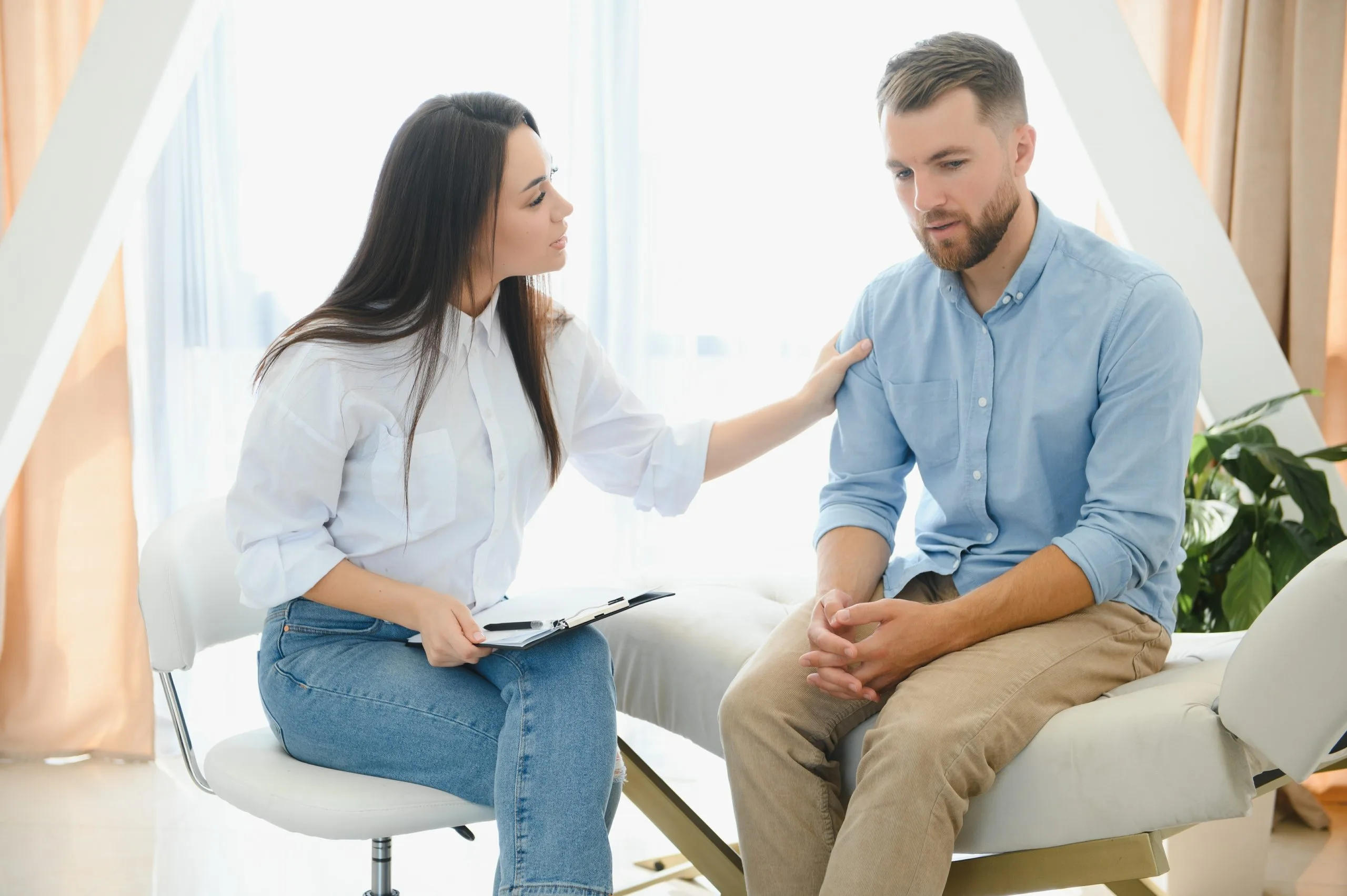 Female therapist with clipboard comforting male patient on couch in bright office