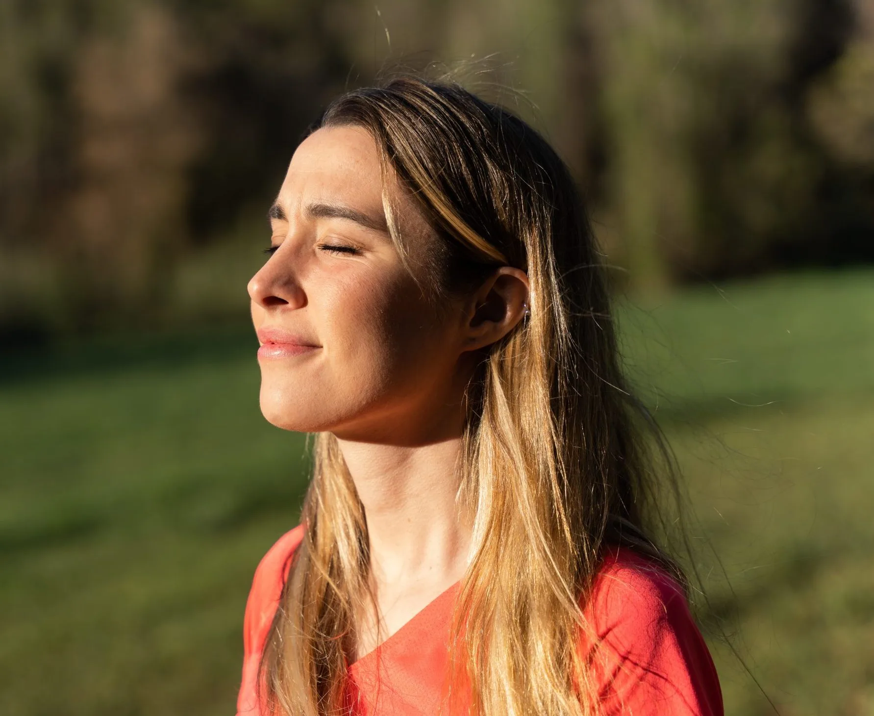 Woman in coral shirt outdoors with eyes closed, natural green background