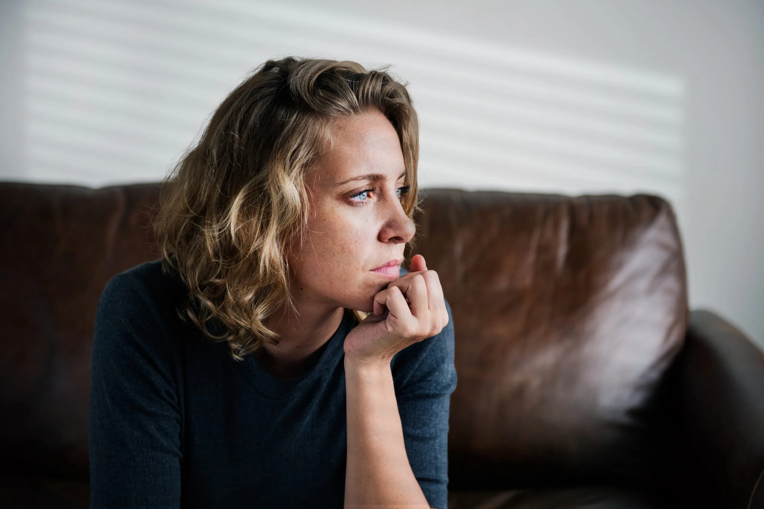 Woman in dark blue shirt sitting on brown couch with hand to face, looking worried