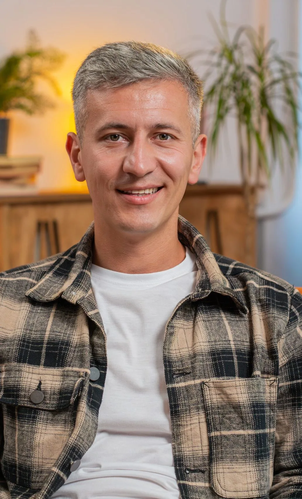 Man in plaid shirt smiling at camera with plants and warm lighting in background