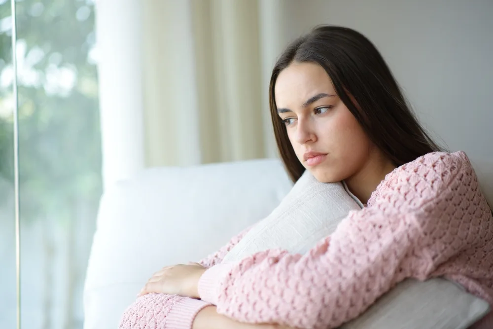 Woman in pink cardigan sitting on white couch looking downward pensively