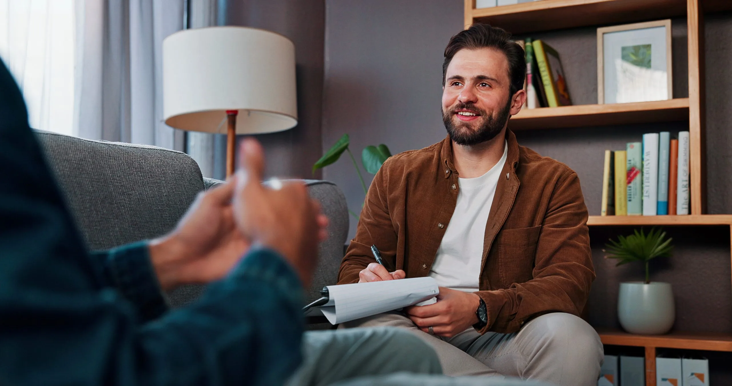 Male therapist taking notes while listening to patient in bright office with bookshelves