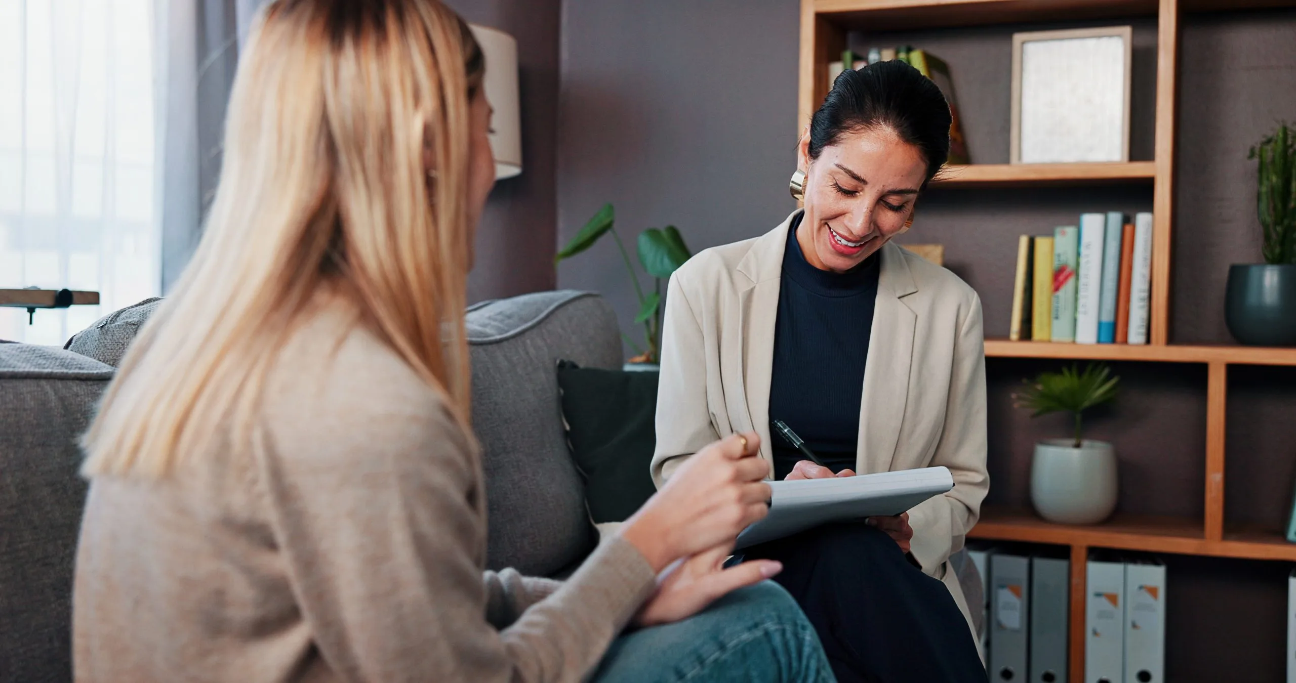 Therapist taking notes on a tablet while listening to a patient on a gray couch with bookshelves behind