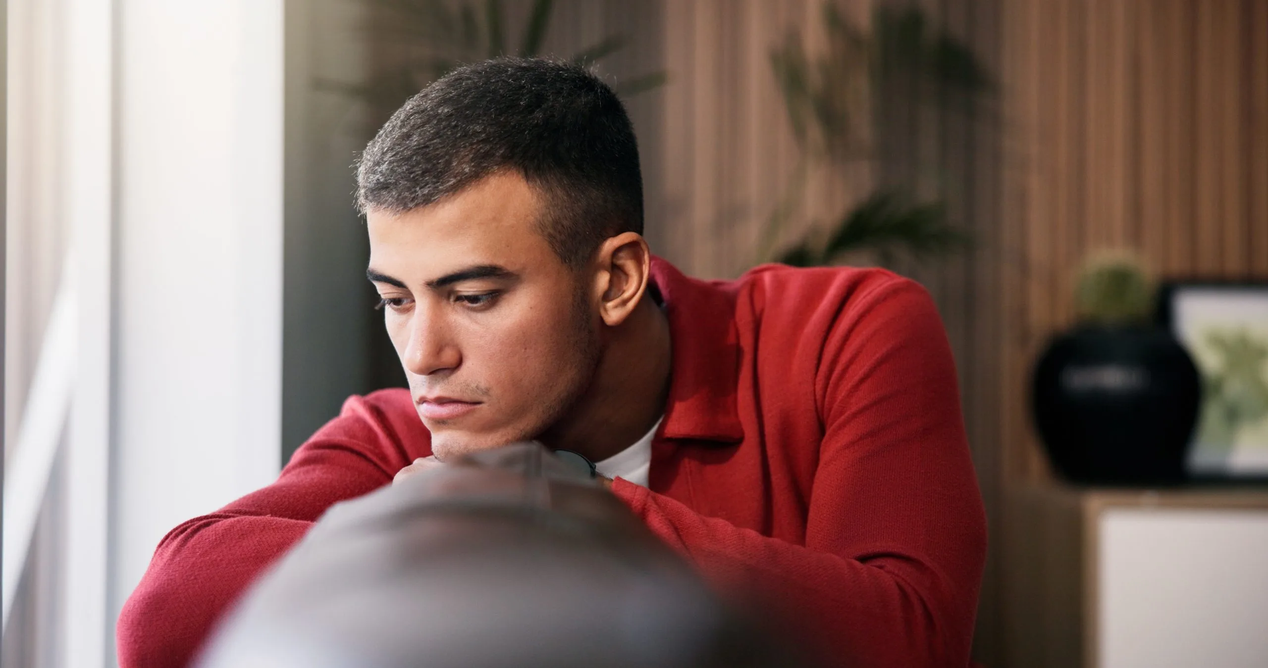 Man in red shirt leaning forward on chair indoors with plants in background