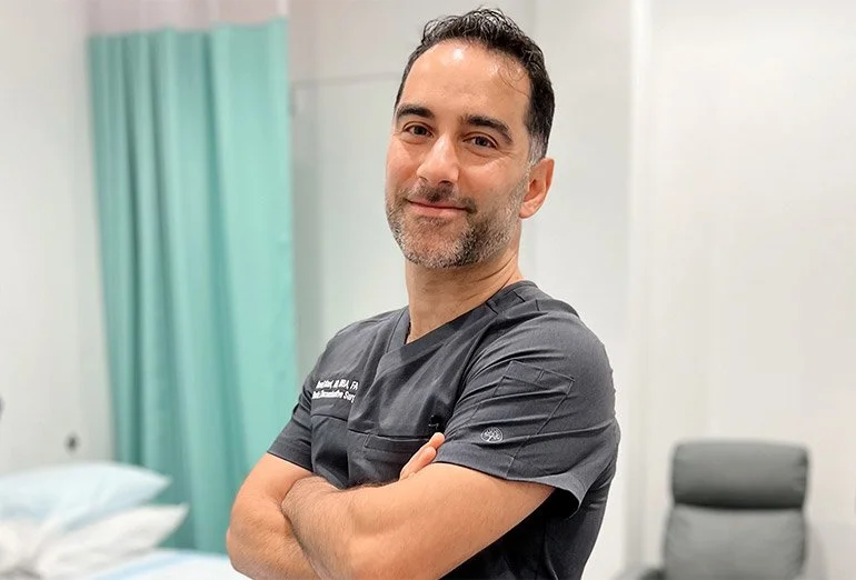 Surgeon in dark scrubs with arms crossed, standing in clinical room with turquoise curtain