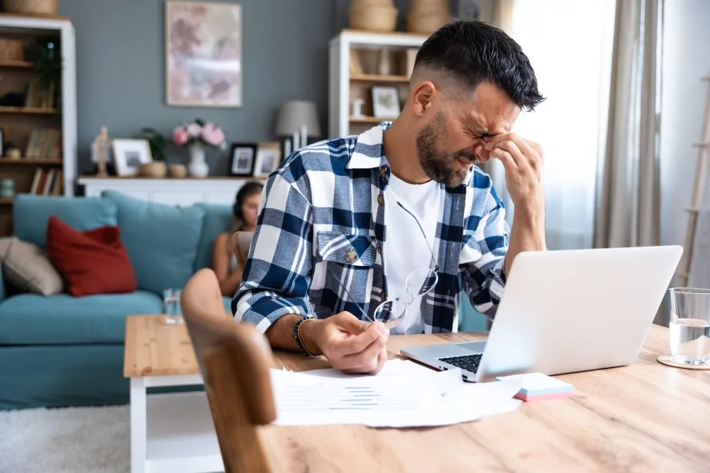 Stressed adult man pinching the bridge of his nose at a laptop, struggling to focus - adult ADHD symptoms