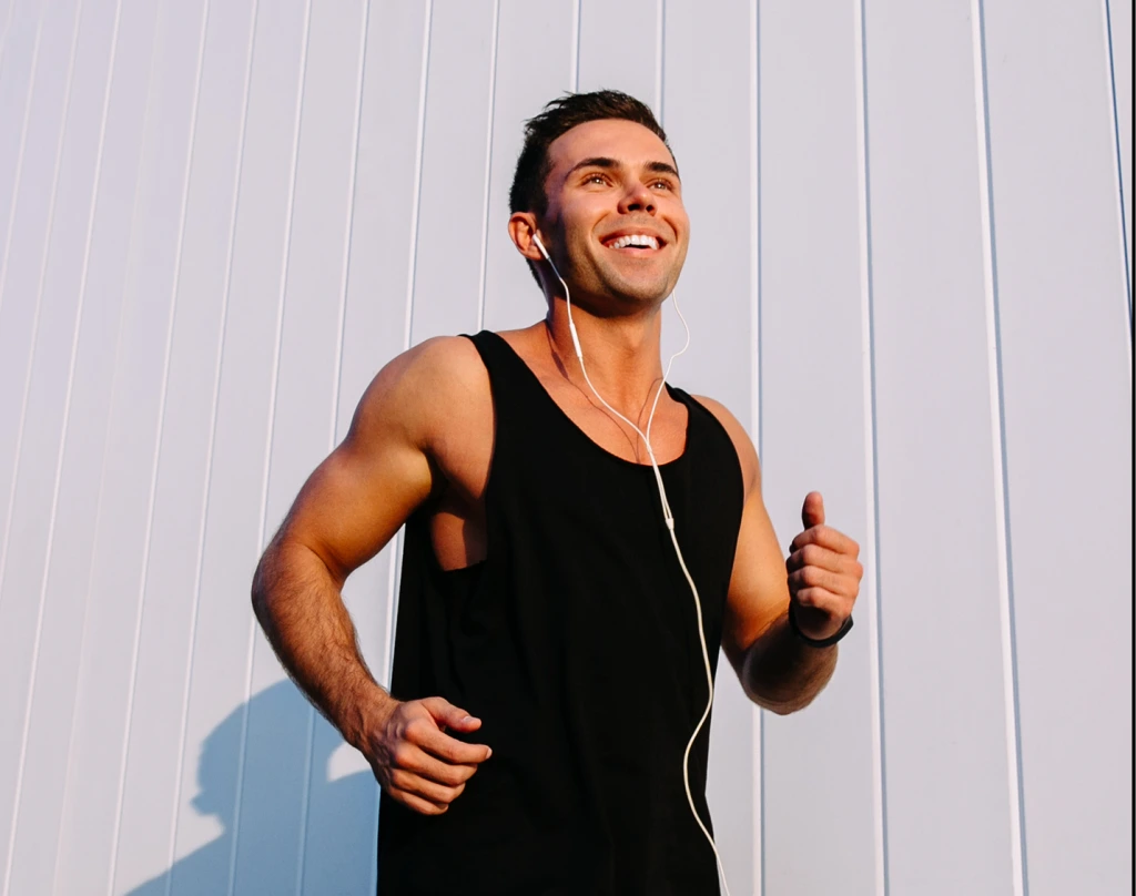 Man jogging outdoors wearing black tank top and white earbuds, smiling while running