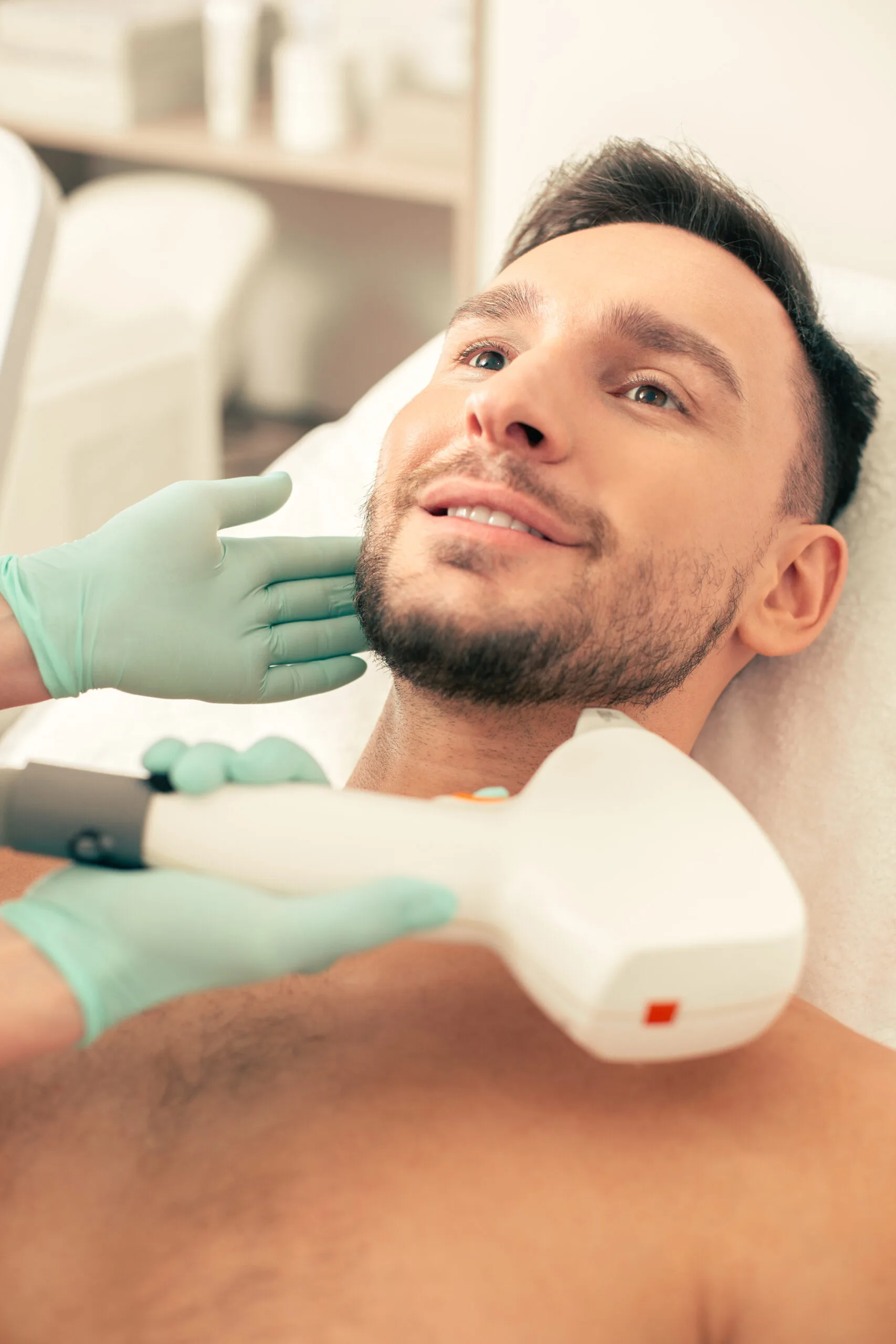 Clinician in teal gloves examining male patient's facial skin with handheld laser device