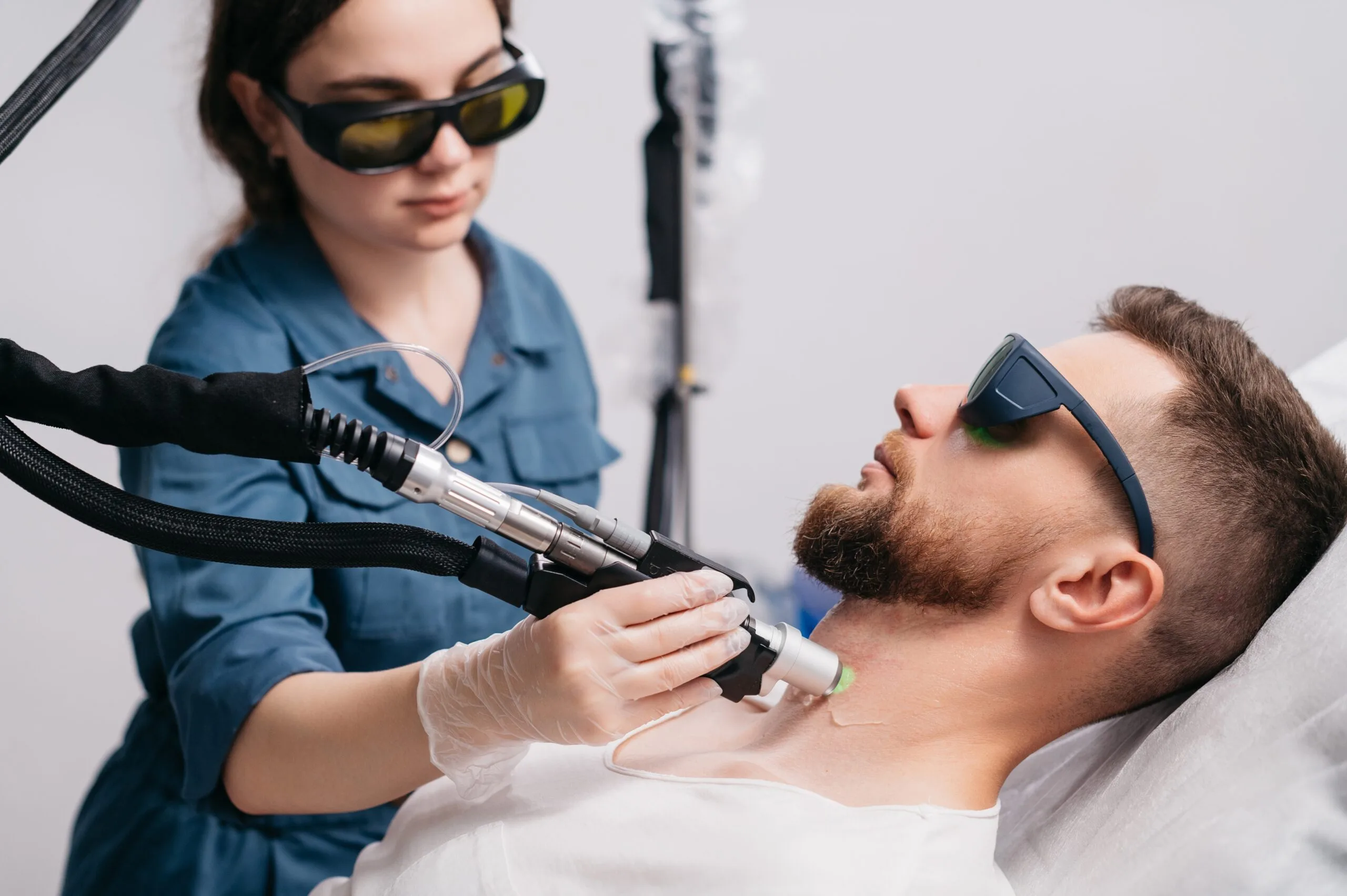 Female technician in protective eyewear using laser handpiece on bearded male patient's face