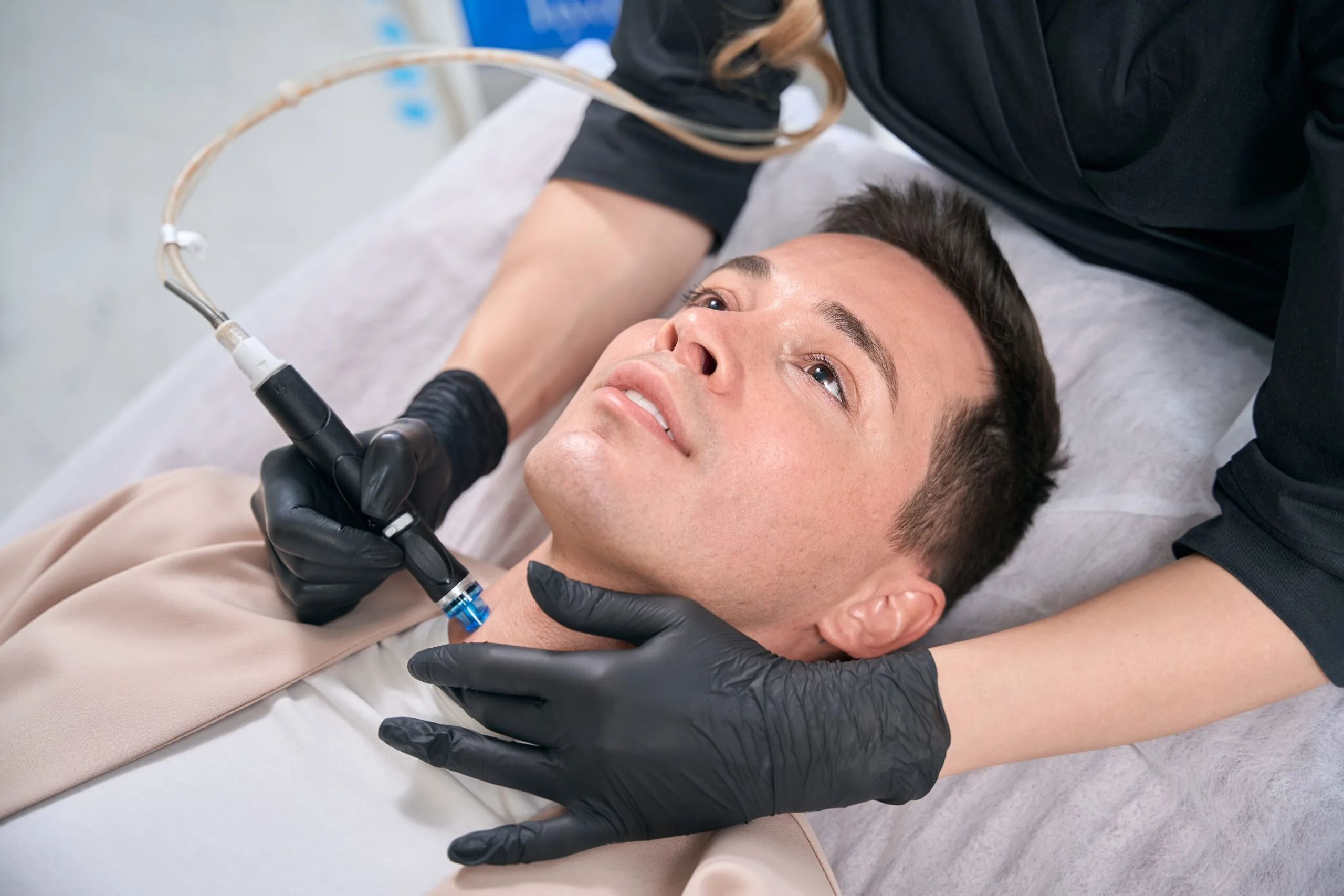 Clinician applying laser treatment to male patient's face lying on treatment bed