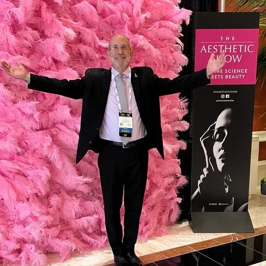 Man in black blazer with arms raised in front of large pink feathered backdrop next to The Aesthetic Now conference signage