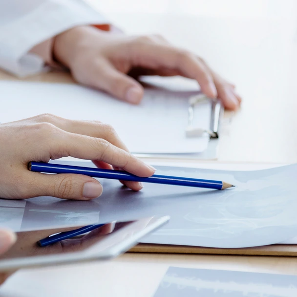 Person holding blue pencil over paperwork on desk