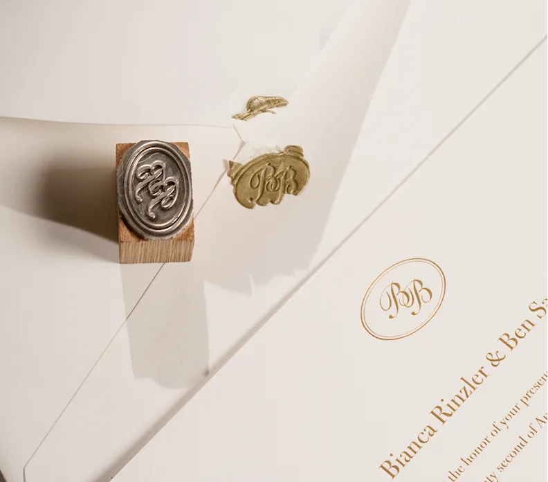 Wooden stamp with monogram, gold foil leaf impressions, and cream stationery with gold oval seal and recipient name